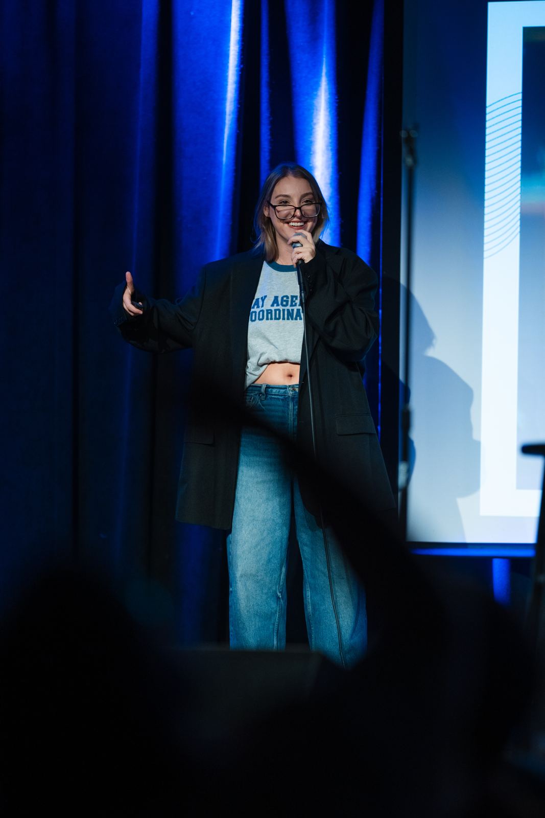 Madison Walser on stage in an oversized black blazer, mic in one hand, the other hand out mid-point